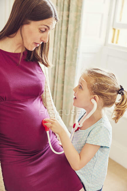 Дівчина з іграшковим стетоскопом слухає шлунок вагітної матері — стокове фото