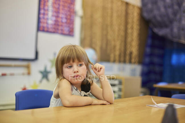 Colegiala primaria jugueteando con el pelo en el escritorio del aula - foto de stock