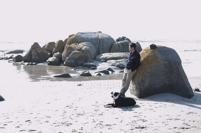 Hombre adulto medio con perro apoyado en la roca mirando al mar, Ciudad del Cabo, Cabo Occidental, Sudáfrica - foto de stock