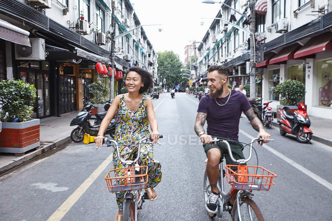 Casal de hipster étnico multi ciclismo ao longo da rua da cidade, Shanghai French Concession, Shanghai, China — Fotografia de Stock