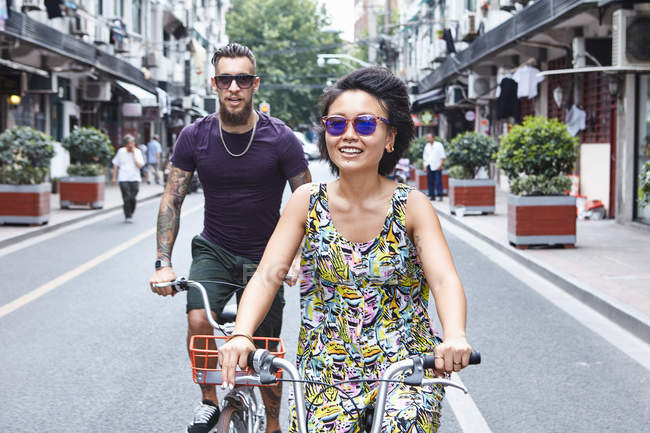 Couple hipster multi ethnique à vélo le long de la rue de la ville, Shanghai concession française, Shanghai, Chine — Photo de stock