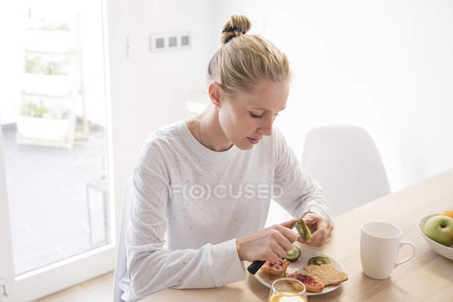 Young woman peeling kiwi at breakfast table — Stock Photo