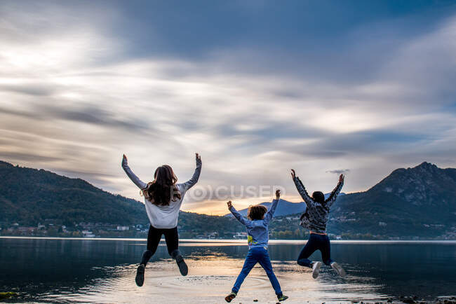 Veduta posteriore del ragazzo e delle giovani donne che saltano in aria lungo il fiume al tramonto, Vercurago, Lombardia, Italia — Foto stock