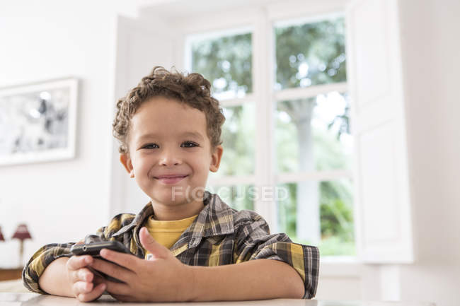Хлопчик за столом тримає мобільний телефон і посміхається на камеру — стокове фото