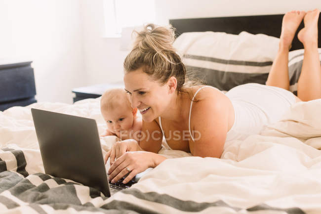 Baby girl and mother lying on bed with laptop — Stock Photo