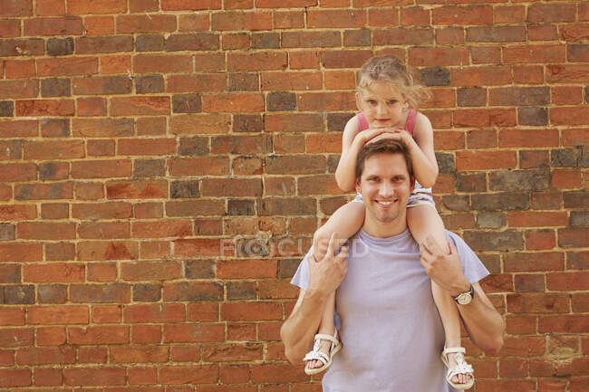 Retrato del hombre adulto medio llevando a la hija en hombros por la pared de ladrillo - foto de stock