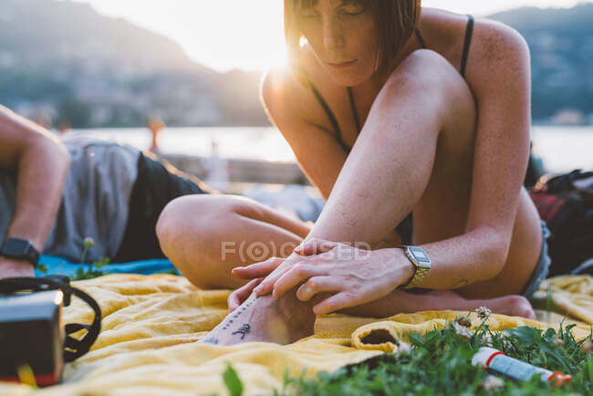 Mujer joven mirando el tatuaje del pie en la hierba frente al mar, Lago de Como, Lombardía, Italia - foto de stock