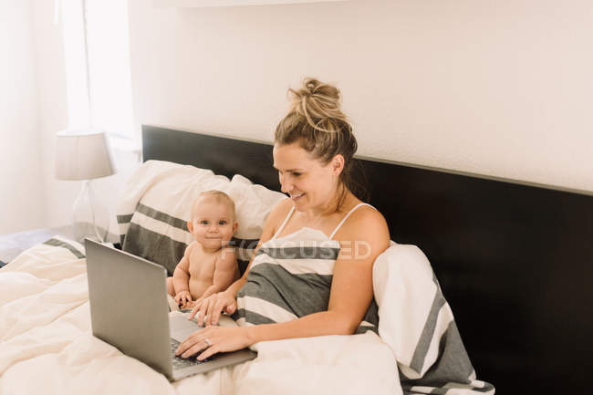 Porträt eines süßen Mädchens, das im Bett sitzt, während die Mutter Laptop benutzt — Stockfoto