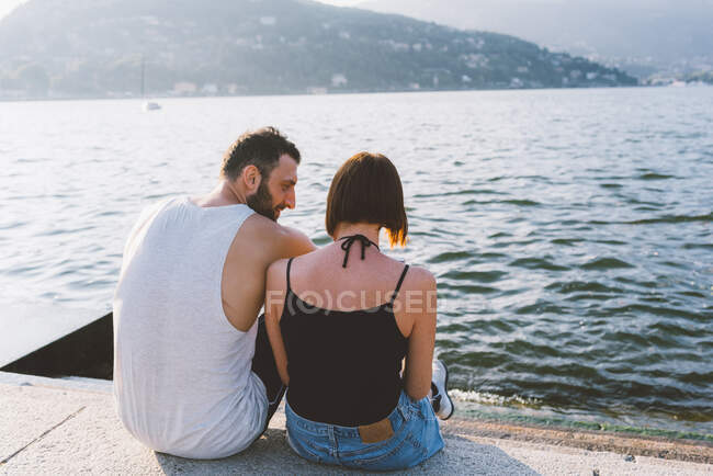 Vista trasera de la joven pareja sentada en el paseo marítimo, Lago de Como, Lombardía, Italia - foto de stock