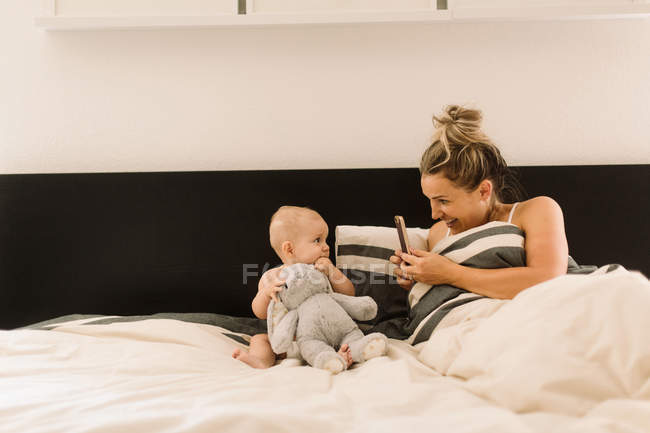 Мама фотографирует маленькую дочь в постели с мягкой игрушкой — стоковое фото