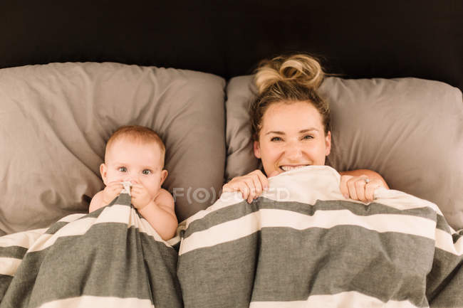 Portrait of woman lying in bed under duvet with baby daughter — Stock Photo