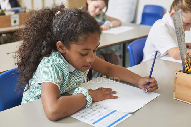 Colegiala escribiendo en el escritorio del aula en la lección de la escuela primaria - foto de stock