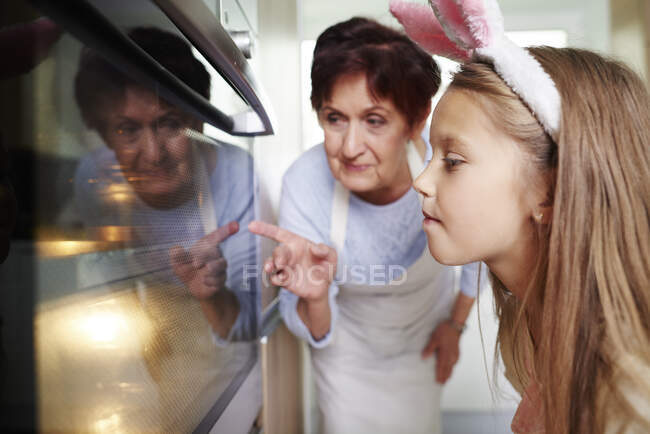 Девочка и бабушка смотрят пасхальное печенье в кухонной печи — стоковое фото