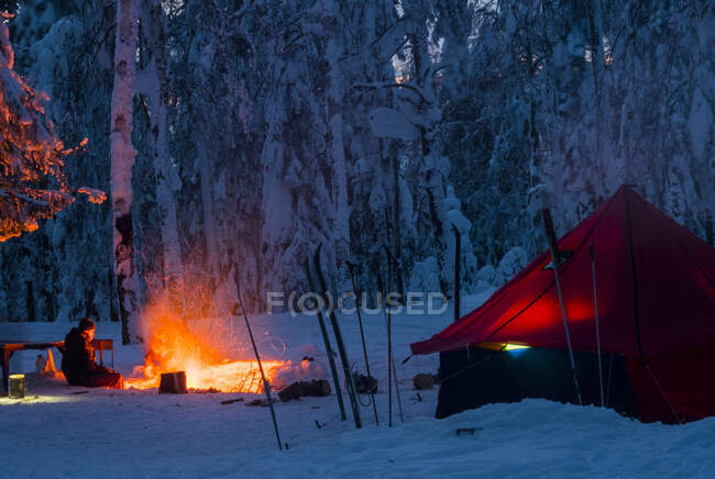 Homme assis à côté du feu de camp, la nuit, près de la tente, dans la forêt enneigée, Russie — Photo de stock