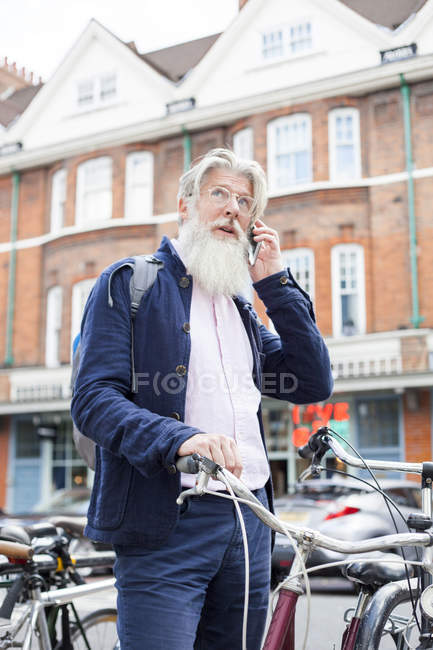 Hombre maduro de pie junto a la bicicleta y hablando en el teléfono inteligente - foto de stock
