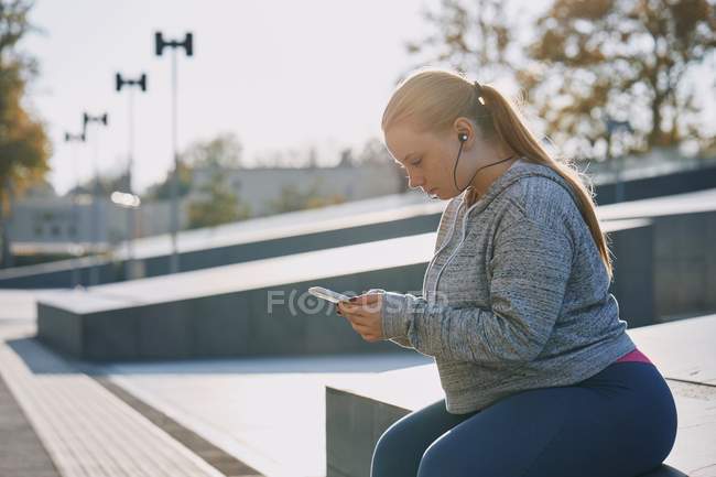 Curvaceous young woman training and sitting on wall with smartphone — Stock Photo