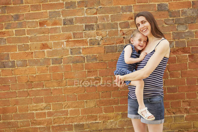 Porträt einer schwangeren mittleren erwachsenen Frau, die ihre Tochter an einer Ziegelwand trägt — Stockfoto