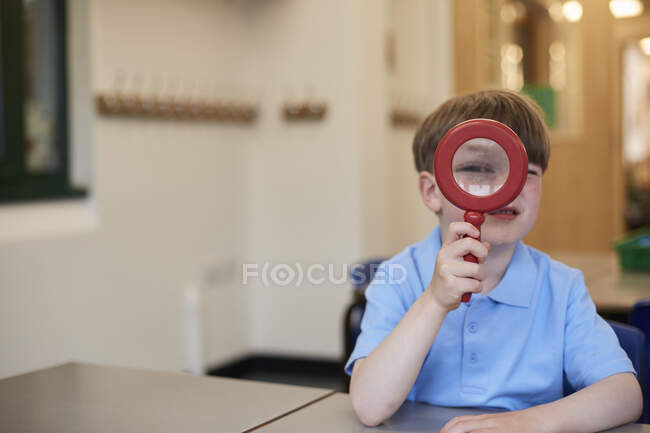 Schoolboy looking through magnifying glass in classroom at primary school, portrait — Stock Photo