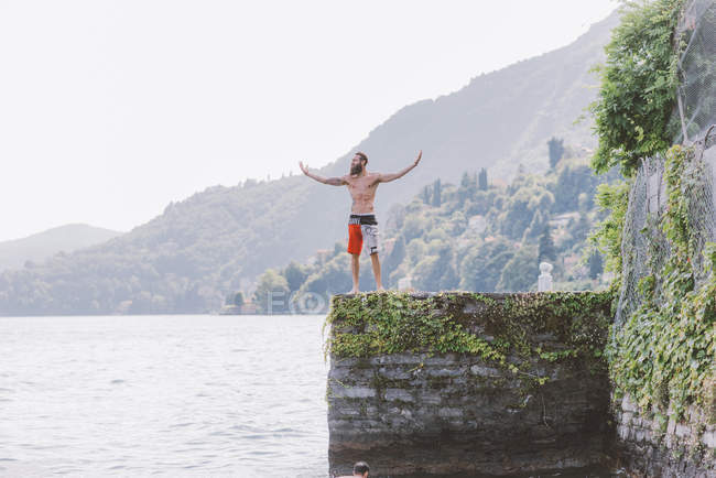 Молодой мужчина стоит на пирсе с распростертыми объятиями, озеро Комо, Ломбардия, Италия — стоковое фото