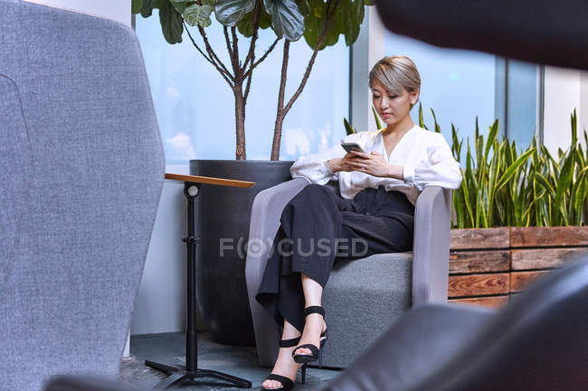 Бізнес-леді сидить на стільці в офісі і використовує смартфон — стокове фото