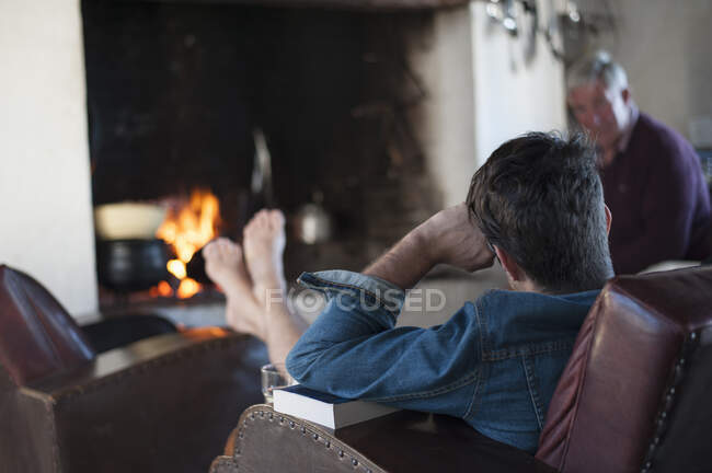Vista por encima del hombro del hombre mayor y el hijo en casa frente al fuego de leña - foto de stock