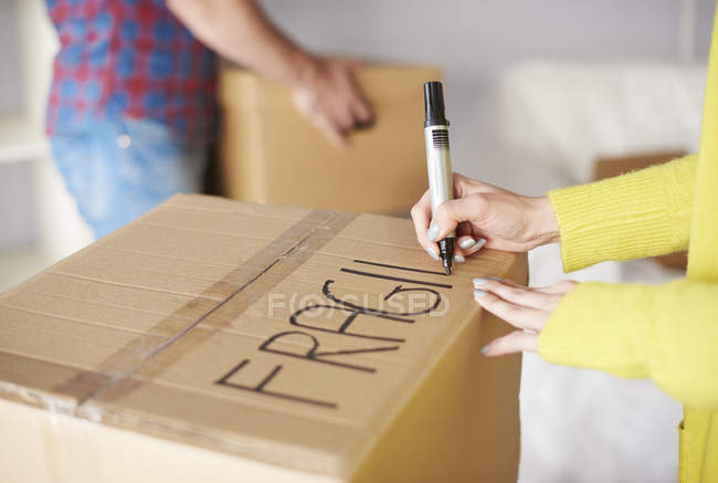 Pareja joven mudándose a casa, mujer joven etiquetando caja de cartón, sección central - foto de stock