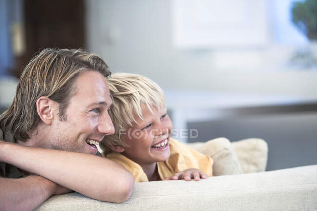 Отец и сын отдыхают на диване, отводя взгляд улыбаясь — стоковое фото