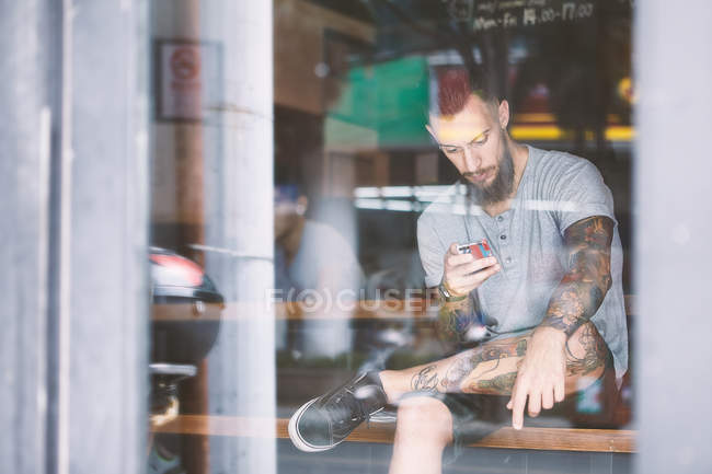 Joven hipster masculino en el asiento de la ventana de la cafetería mirando el teléfono inteligente, Shanghai French Concession, Shanghai, China - foto de stock