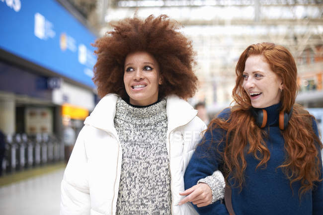 Dos mujeres jóvenes en la estación de tren caminando brazo en brazo - foto de stock