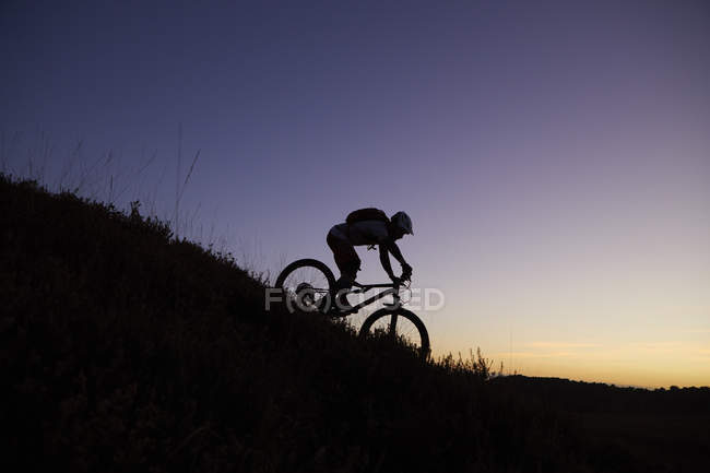 Silueta de ciclista de montaña macho montando en colina abajo al atardecer - foto de stock