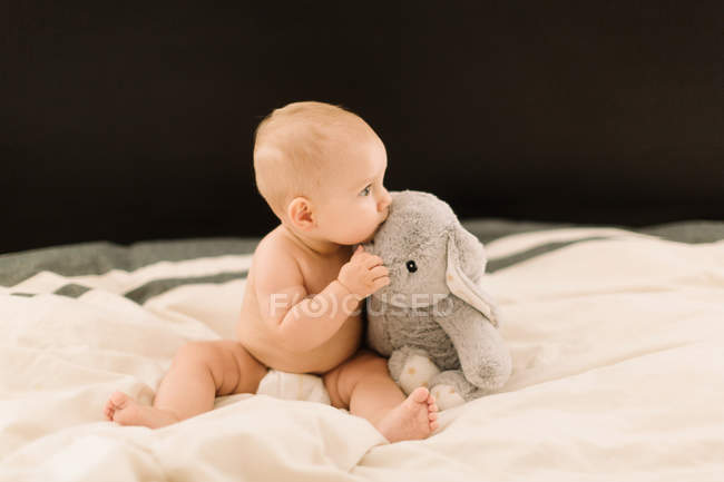 Cute baby girl sitting on bed with soft toy — Stock Photo