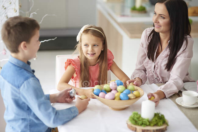 Дівчина з братом і матір'ю готує барвисті великодні яйця за обіднім столом — стокове фото