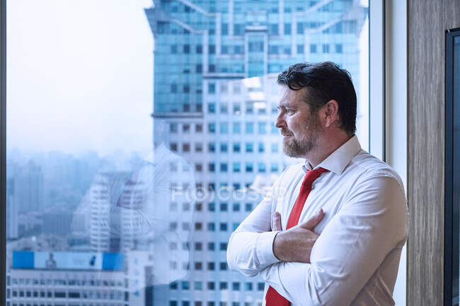 Businessman, arms crossed, looking through window at cityscape — Stock Photo