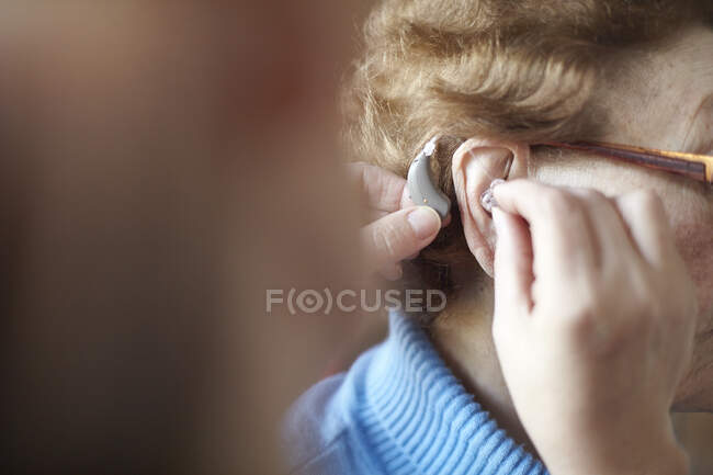 Femme mature aidant une femme âgée à insérer une aide auditive, un gros plan, une mise au point différentielle — Photo de stock