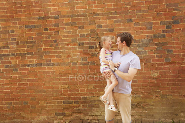 Chica riendo en brazos de padre por pared de ladrillo - foto de stock