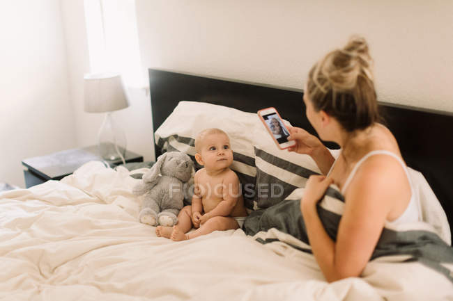 Mãe tirando foto de bebê filha sentada na cama com brinquedo macio — Fotografia de Stock