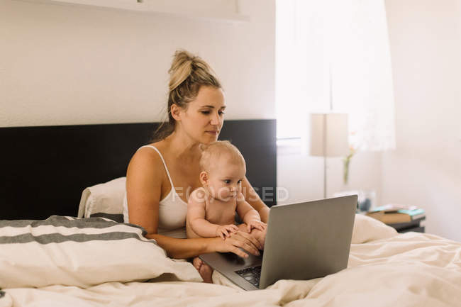 Bebê menina e mãe sentado na cama com laptop — Fotografia de Stock