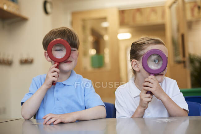 Schoolboy and girl looking through magnifying glasses in classroom at primary school, portrait — Stock Photo