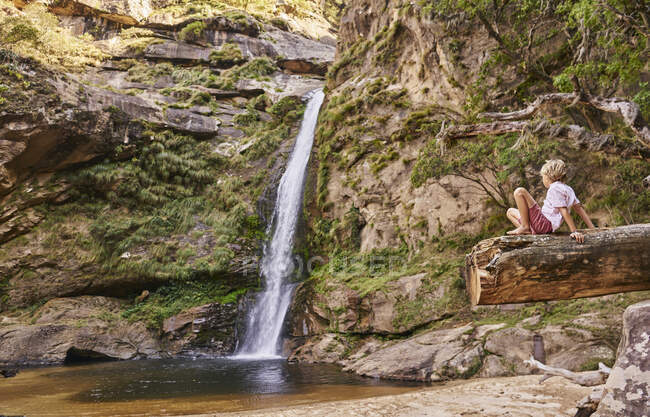 Niño sentado en el tronco mirando cascada, Samaipata, Santa Cruz, Bolivia, América del Sur - foto de stock