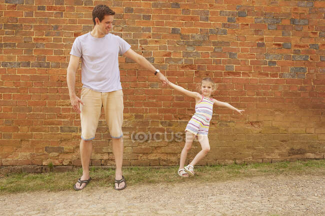 Девушка наклоняется боком, держа отца за руку за кирпичную стену — стоковое фото
