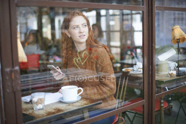 Перегляд через вікно жінки в кав'ярні тримає мобільний телефон — стокове фото