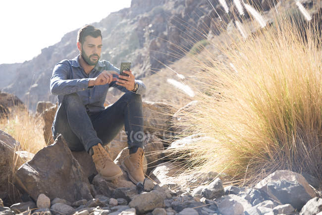 Junge männliche Wanderer sitzen auf Felsbrocken und schauen auf ihr Smartphone im Tal, Las Palmas, Kanarische Inseln, Spanien — Stockfoto