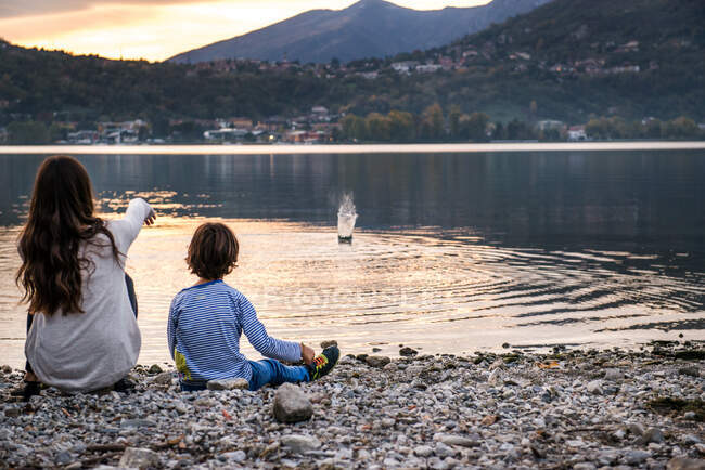 Rear view of boy and young woman throwing pebbles into river at dusk, Vercurago, Lombardy, Italy — Stock Photo