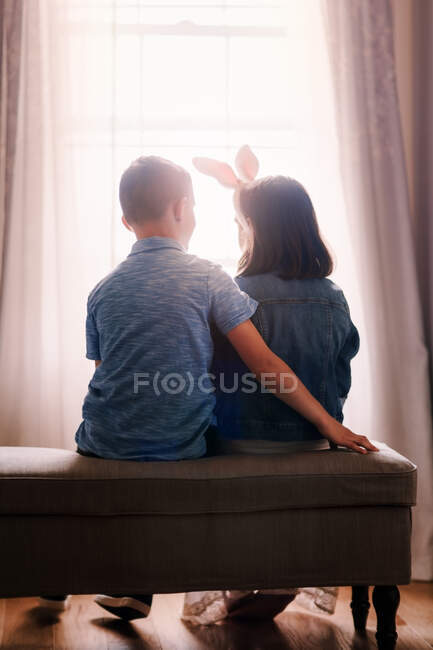 Niño y niña, sentados frente a la ventana, niña con orejas de conejo, vista trasera - foto de stock