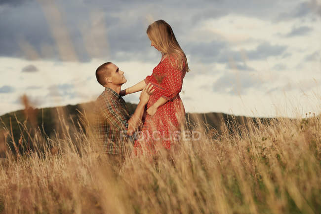 Чоловік середнього віку на колінах з рукою на животі вагітної дружини в полі — стокове фото