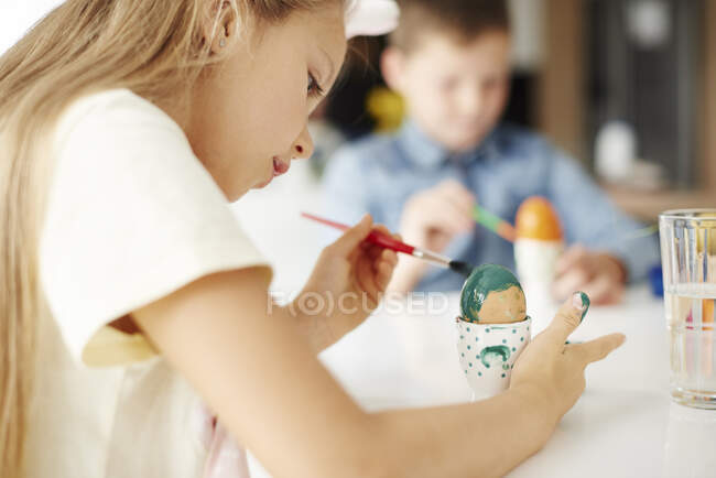 Девушка рисует варёное пасхальное яйцо зеленым за столом — стоковое фото