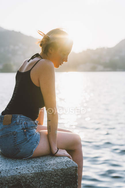 Mujer joven sentada en la pared frente al mar en el Lago de Como, Lombardía, Italia - foto de stock