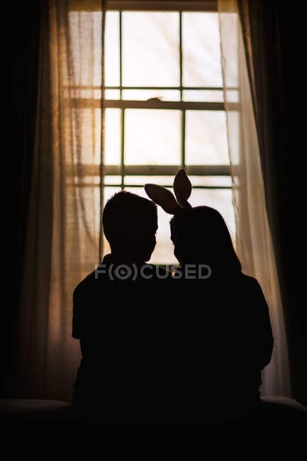 Silhueta de menino e menina, sentado na frente da janela, menina vestindo orelhas de coelho, visão traseira — Fotografia de Stock