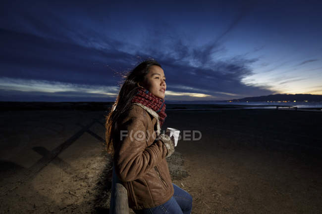 Jovem envolta em cachecol olhando para fora da praia ao entardecer — Fotografia de Stock