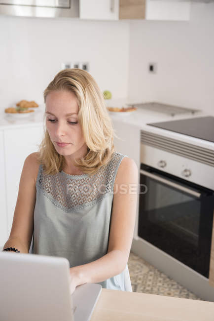 Giovane donna digitando sul computer portatile al tavolo della cucina — Foto stock
